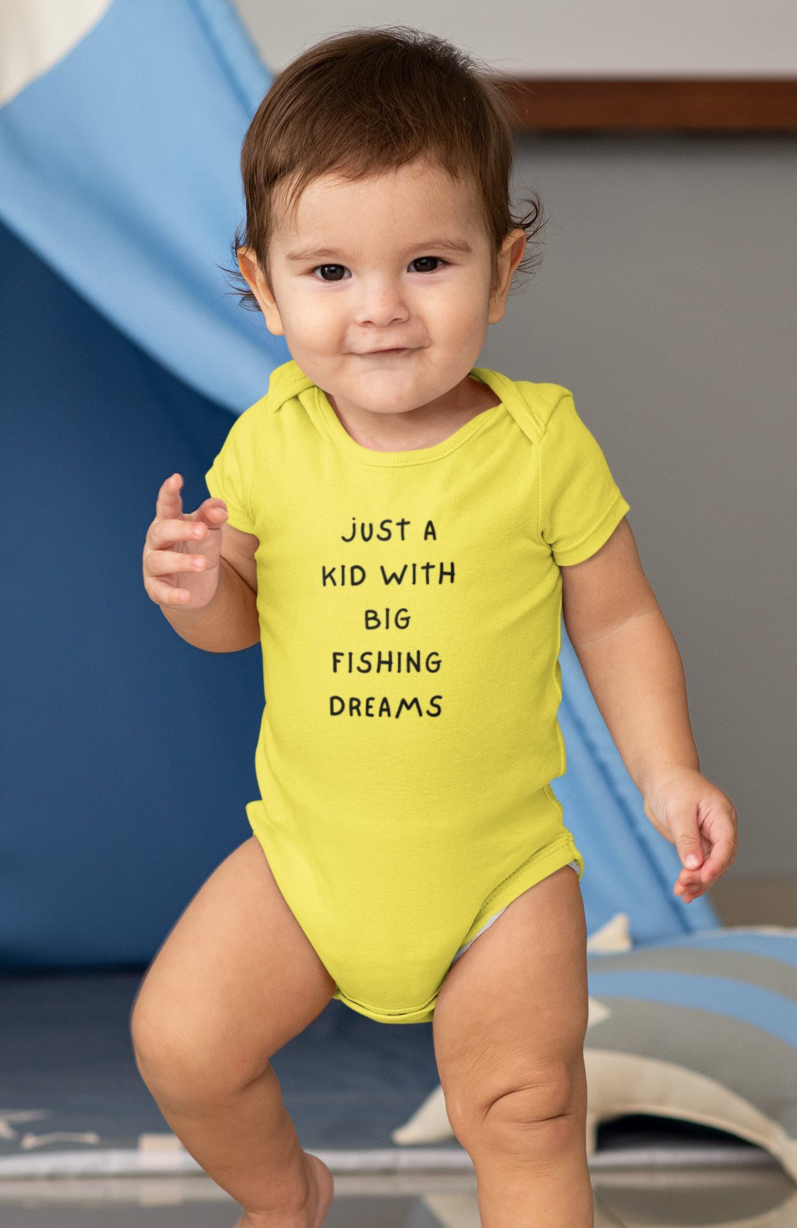 Baby wearing a yellow onesie with text, standing in front of a blue slide.