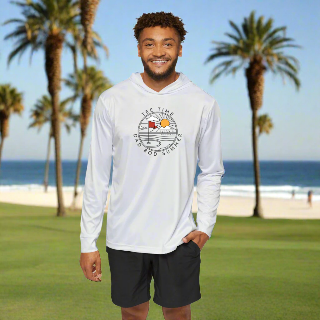 A man wearing a white long sleeve golf hoodie with a graphic print in the center, standing on a golf course with palm trees in the background and playing golf in San Diego California.